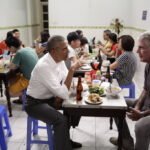 Barack Obama and Anthony Bourdain sitting on plastic stools at a table of bún chả in a Hanoi restaurant